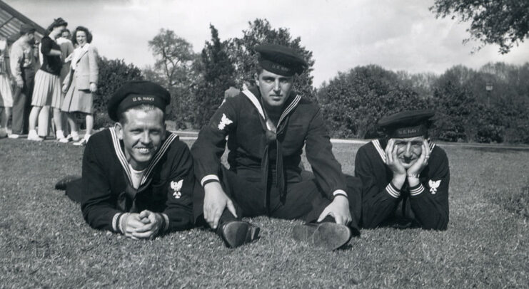 Three US Navy sailors lying in the grass with a group of women standing in the background