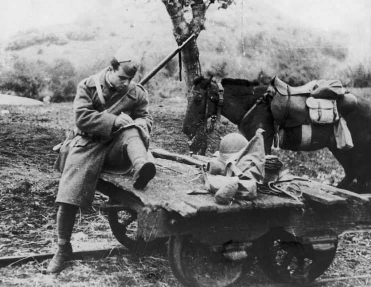 Greek soldier writing a letter in the middle of a muddy road, while his horse stands off to the side