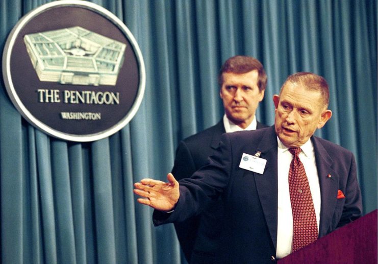 John Singlaub standing behind a podium with William Cohen looking on