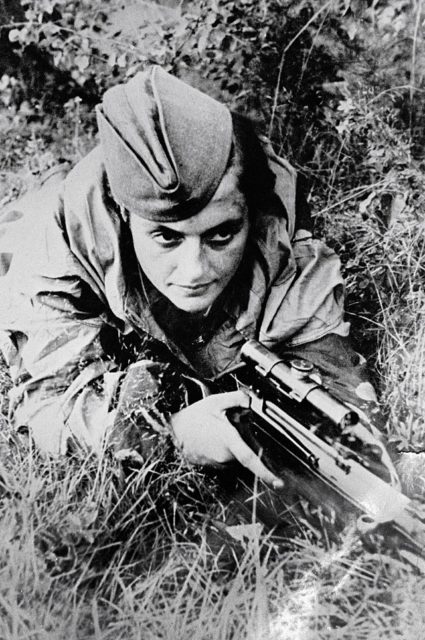 Lyudmila Pavlichenko holding a sniper rifle while lying in tall grass