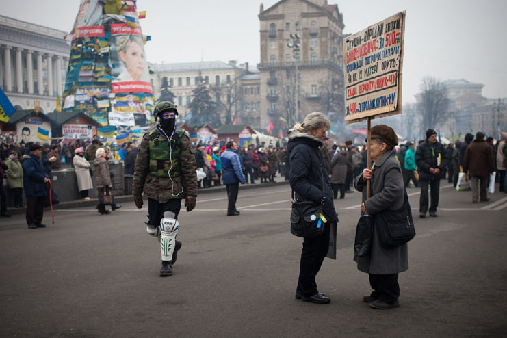 Man dressed in Army clothes walks by two women holding a protest sign