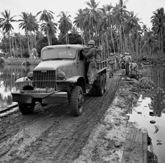 US Army troops driving and walking along a dirt road, surrounded on both sides by water