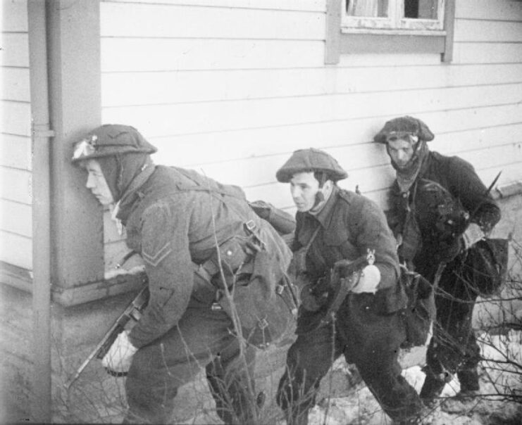 Three British Commandos sneaking along the side of a building