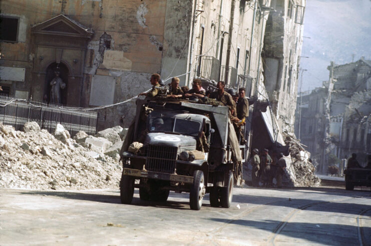 Soldiers riding in a vehicle driving along a road in the middle of a destroyed city
