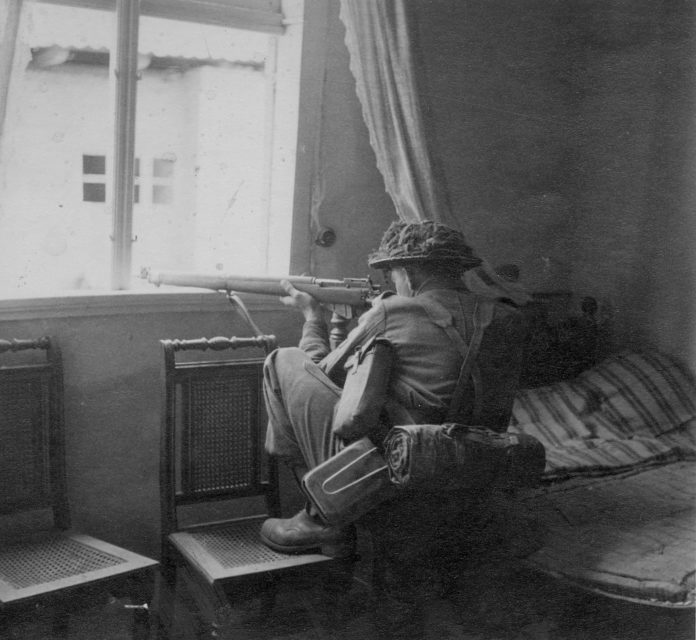 British sniper aiming his weapon out of a bedroom window