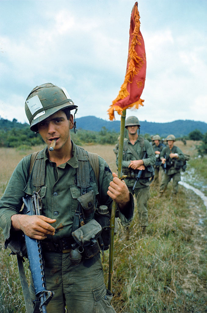 Michael Marrone carrying a captured Viet Cong flag, while his comrades walk single-file behind him