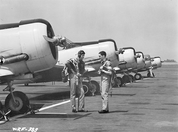 Two pilots stand in front of a row of North American Harvard IIs