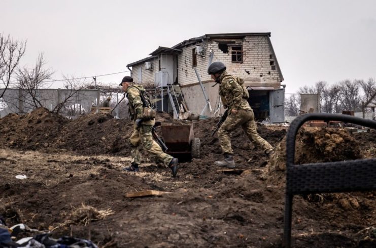 Two Ukrainian soldiers running past a ruined house