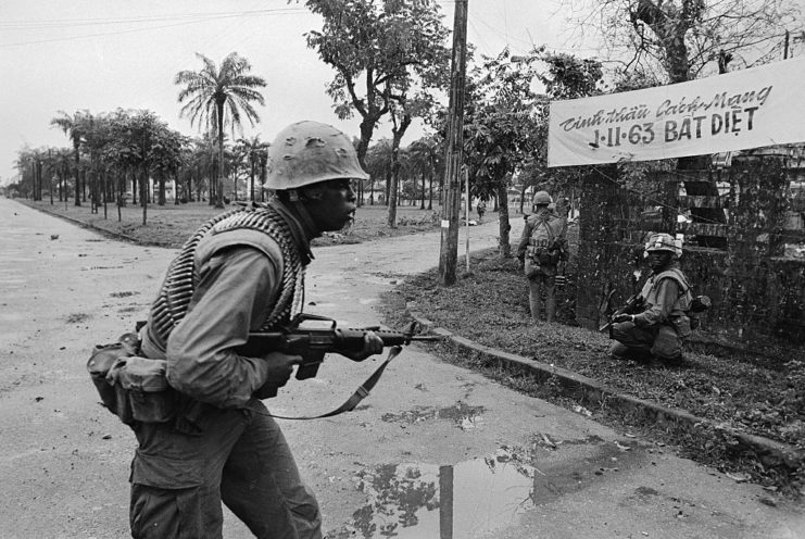 US Marine walking in the middle of a street with a machine gun while two others hide behind a wooden fence