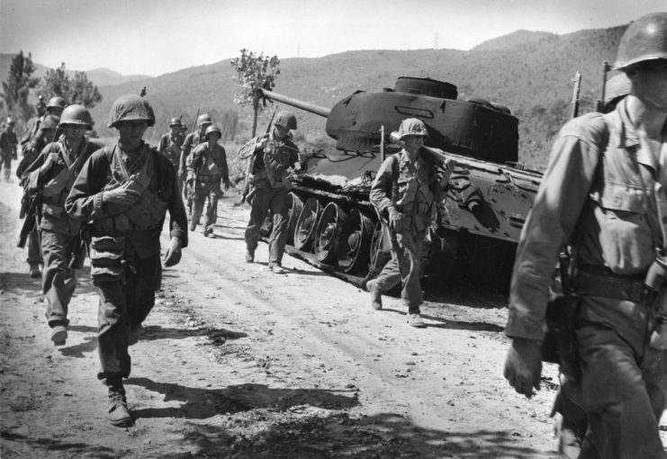 US troops walking past a tank parked on the side of a dirt road