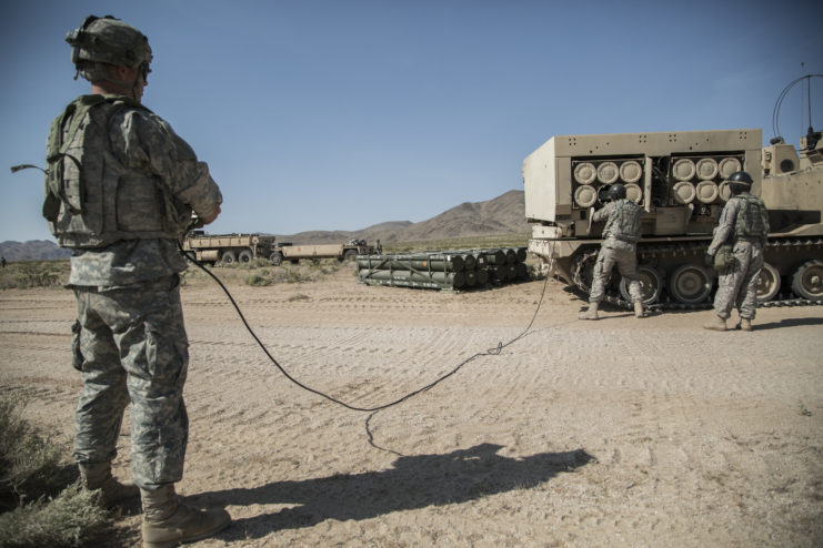 Soldier holding a cord connected to an M270 MLRS while two others load rockets into it