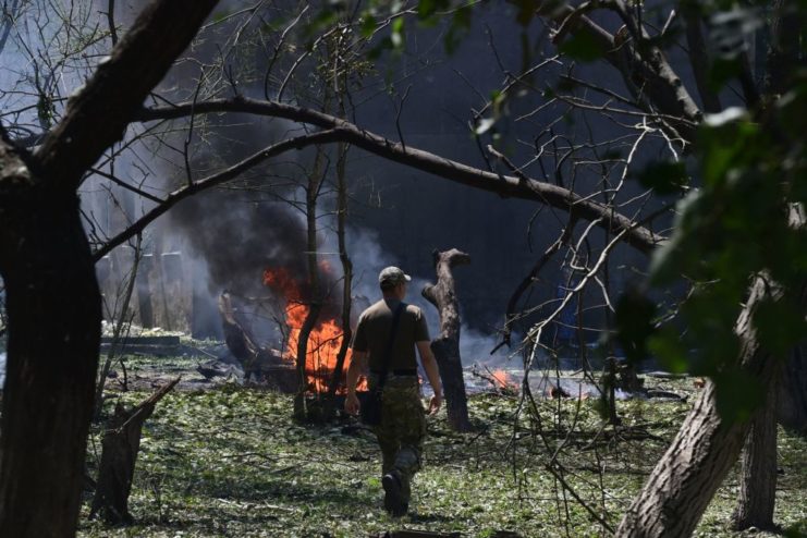 Ukrainian serviceman walking near a burning car shrouded in smoke