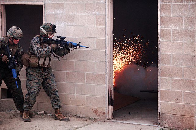 Two US Marines aim their weapons at an open doorway while an explosion occurs inside the building