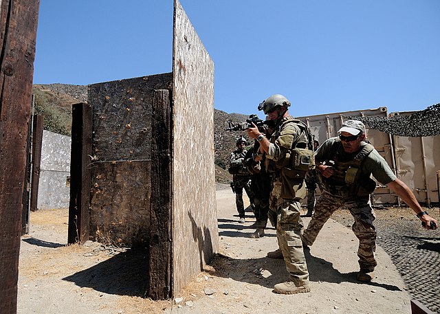 Mass Communication Specialist 1st Class James E. Foehl aiming his gun while Max Joseph prepares to toss a flashbang grenade