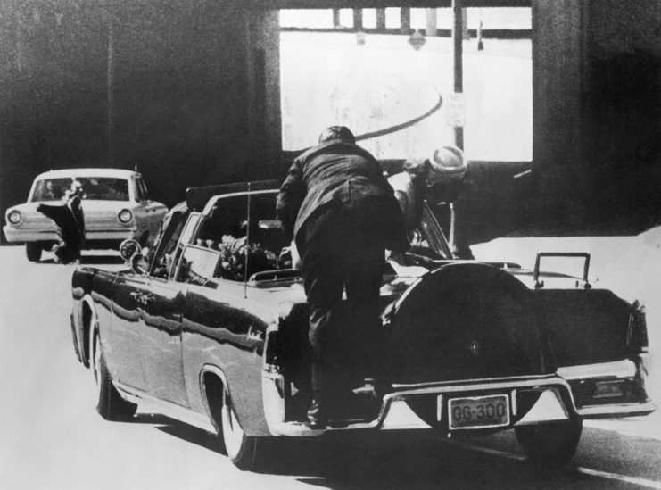 Secret Service agent climbing over the back of a car, while Jacqueline Kennedy leans over John F. Kennedy in the backseat