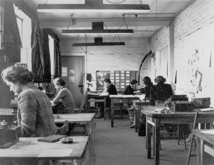 Women leaning over documents at desks set up around a room