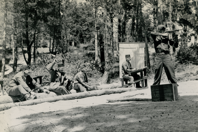 Soldier aiming an M3A1 "Grease Gun" while others watch from behind