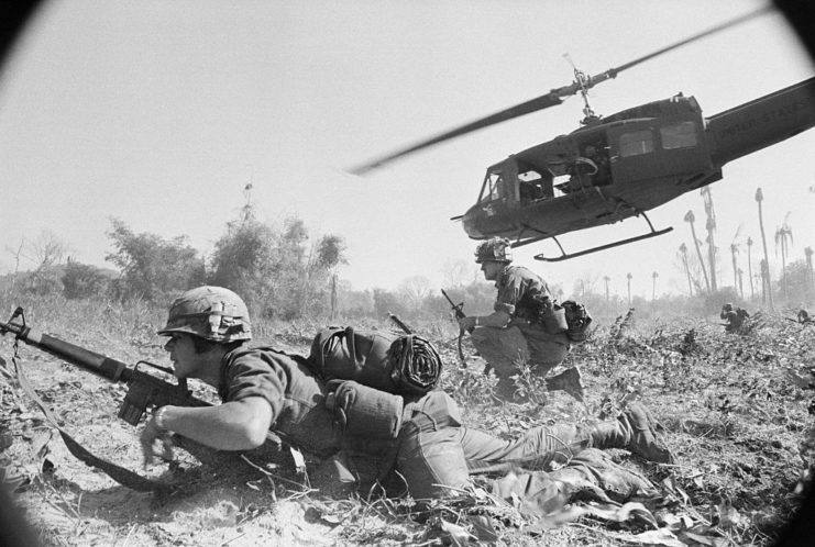 Two US soldiers standing beneath a helicopter hovering over the ground