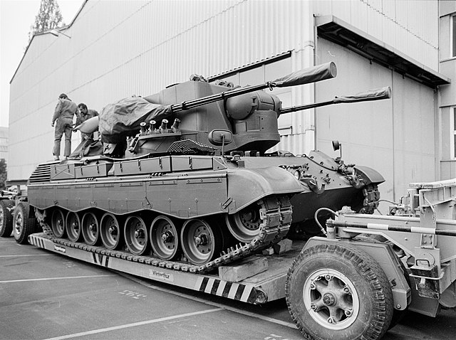 Two individuals standing on a Flakpanzer Gepard parked on the back of a vehicle