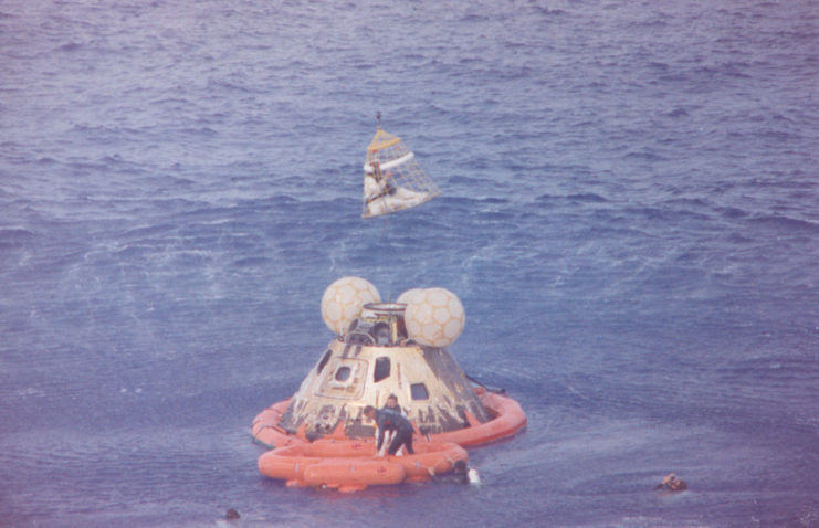 Frogmen swimming around a raft James Lovell Jr. is standing in, while John Swigert Jr. is being lifted into the air