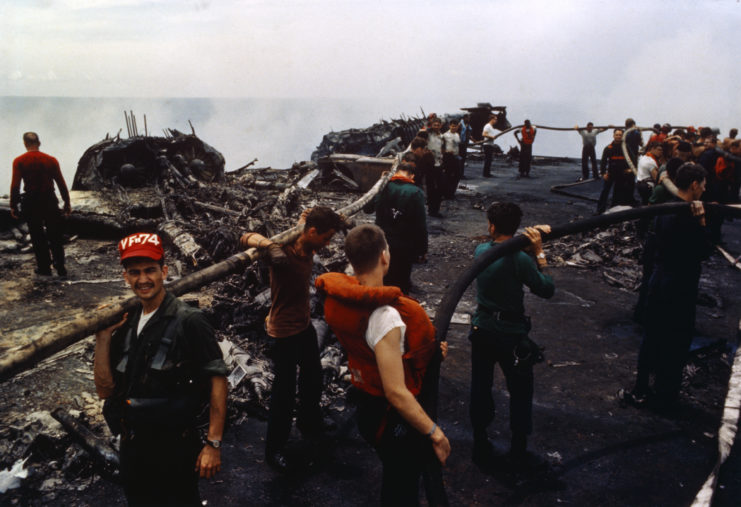 Crewmen carrying hoses on their shoulders amid the smouldering remains on the deck of the USS Forrestal (CV-59)