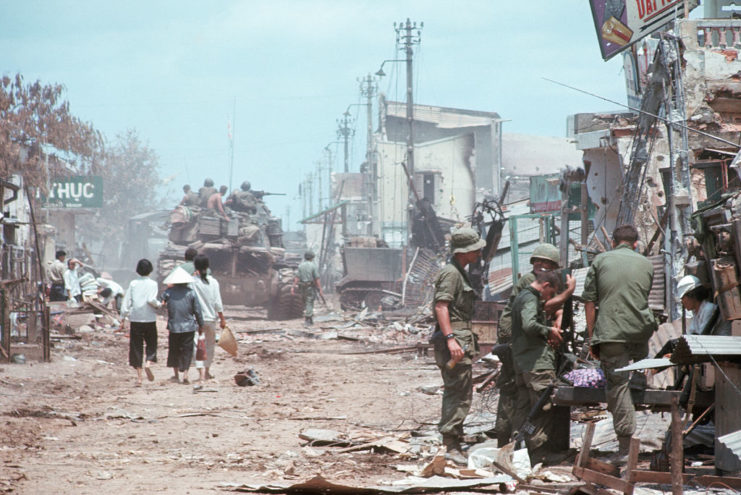 US soldiers, a tank and Vietnamese citizens traveling along a street covered in rubble