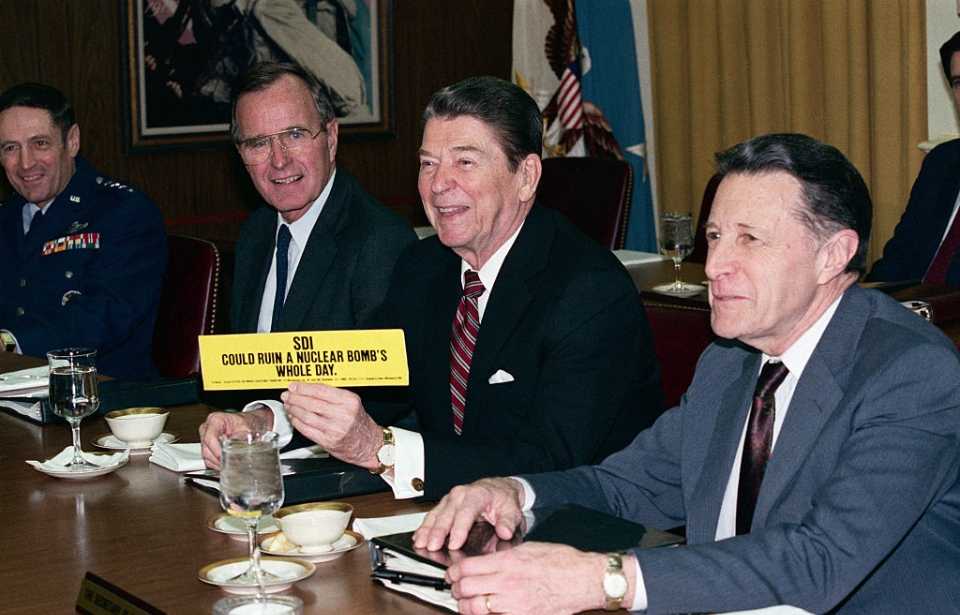 Ronald Reagan sitting at a table with other men while holding a yellow bumper sticker that reads, "SDI could ruin a nuclear bomb's whole day"