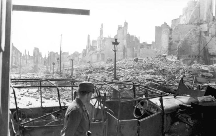 German soldier walking by a destroyed vehicle in a city that's been heavily bombed