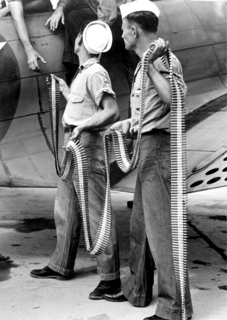 Two ordnancemen handing belted cartridges to another sitting atop a Douglas SBD-3 Dauntless