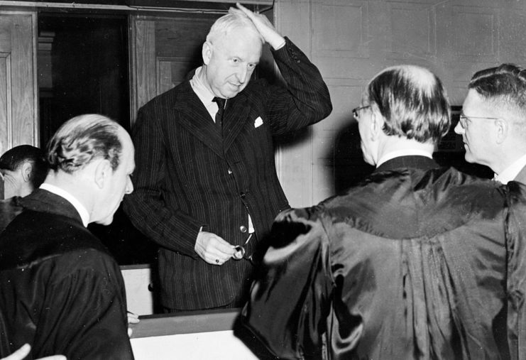 Field Marshal Erich von Manstein standing with judges in robes around him, while slicking back his hair.