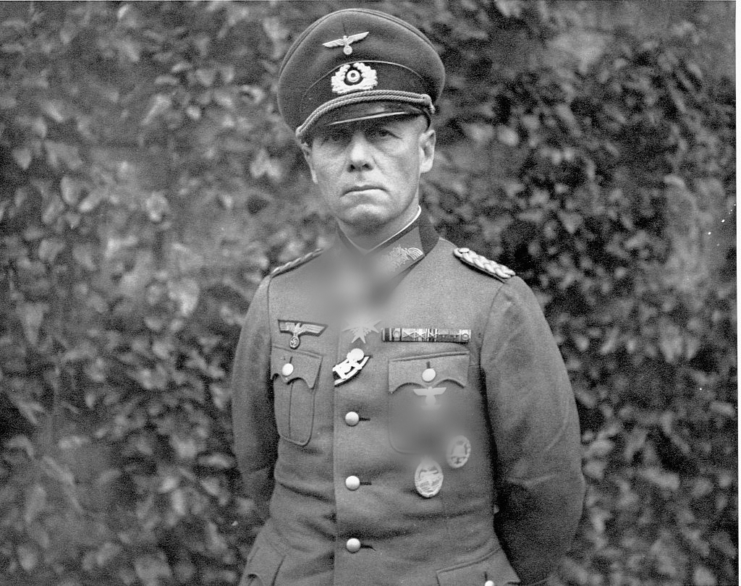 Erwin Rommel in military dress with his hands behind his back in front of a bush.