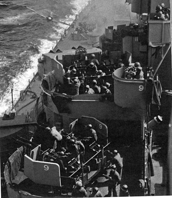 Sailors gathered around the USS Missouri's (BB-63) deck armaments as a kamikaze aircraft flies toward the battleship