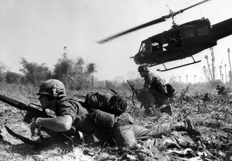 US infantrymen crawling on the ground while a Bell UH-1D Iroquois hovers overhead