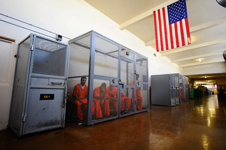 Prisoners sitting in a metal cage within a larger hallway
