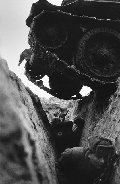 Soldiers hunker down in a trench while a tank rolls over top