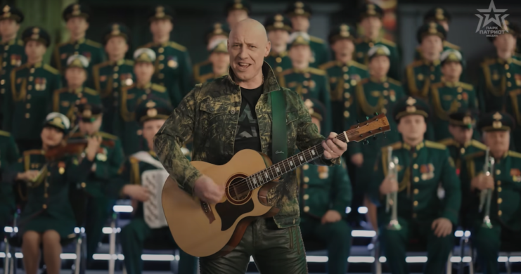 Denis Maidanov playing guitar in front of an orchestra consisting of members of the Russian military