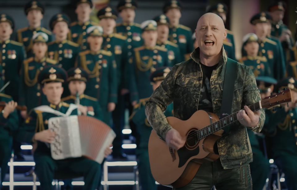 Denis Maidanov playing guitar in front of an orchestra made up of Russian military members