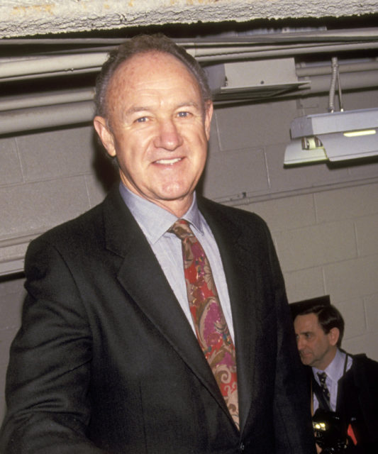 Gene Hackman standing in a low-ceiling room, with a man sitting behind him