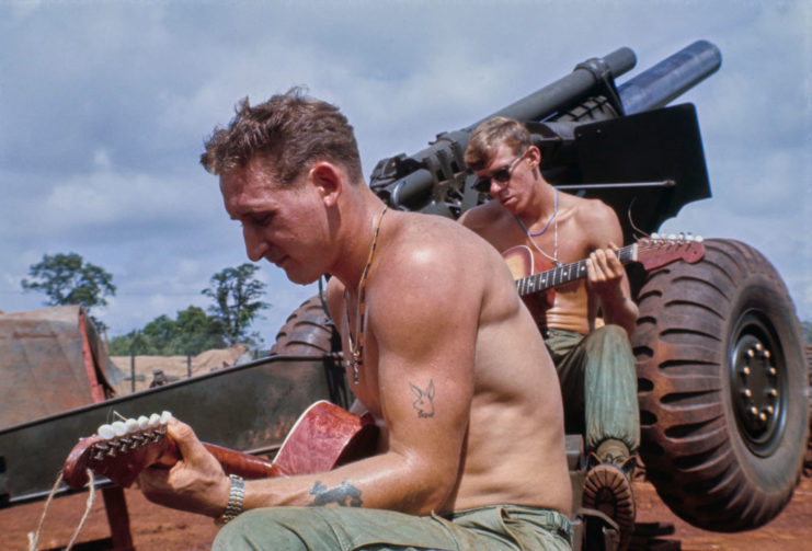 Two members of the 11th Armored Cavalry Regiment playing guitars while sitting on a howitzer