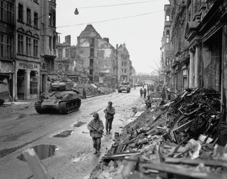 M4A1 Shermans and members of the 3rd Armored Division walking along a street in Cologne