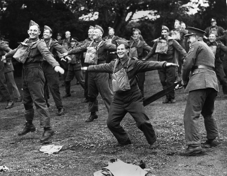 Soldiers with their arms out, while their commander hits one with a plank