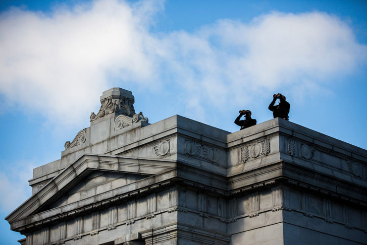 Two Secret Service snipers looking through binoculars while standing on a roof