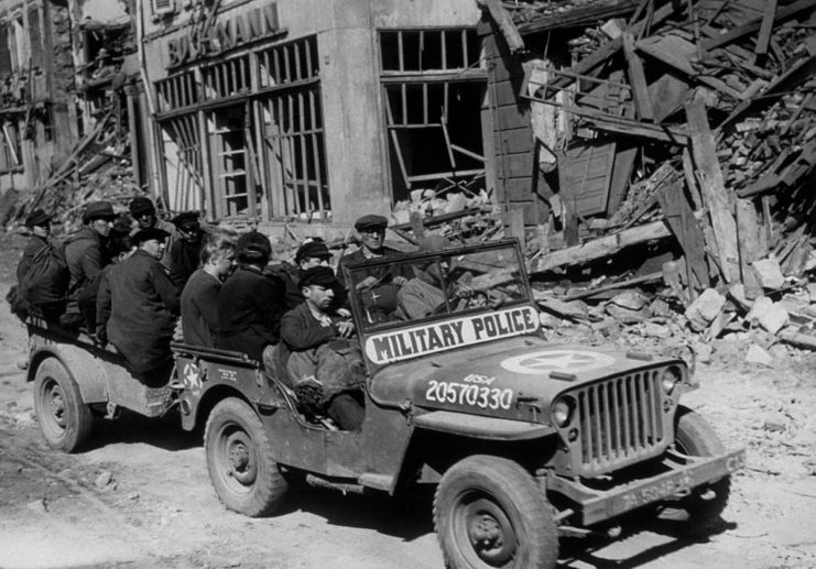 Military police and German civilians riding in a Jeep down a debris-strewn street