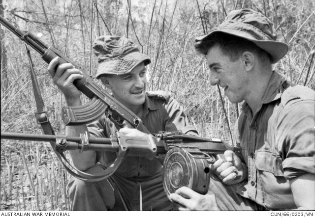 Two Australian soldiers standing together, one holding an AK-47 and the other examining an RPD machine gun