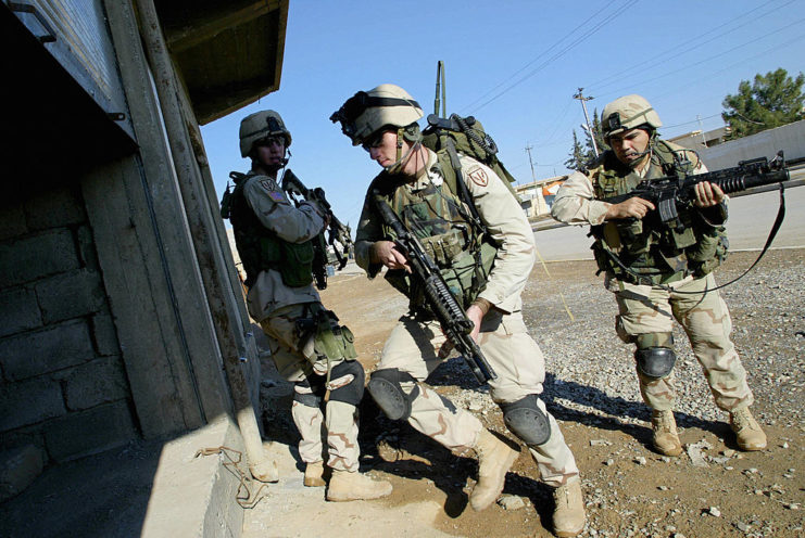 Three American soldiers standing outside of a building while holding their weapons