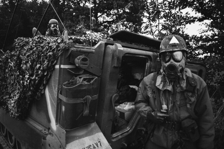 NATO troops standing around a camouflaged tank while wearing gas masks