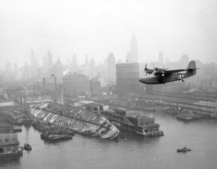 Grumman J4F Widgeon flying past the capsized USS Lafayette (AP-53)