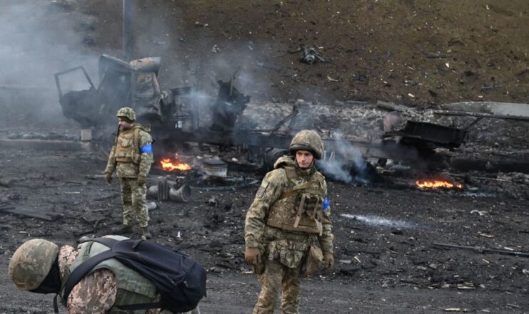 Three Ukrainian soldiers standing among the smouldering remains of a vehicle