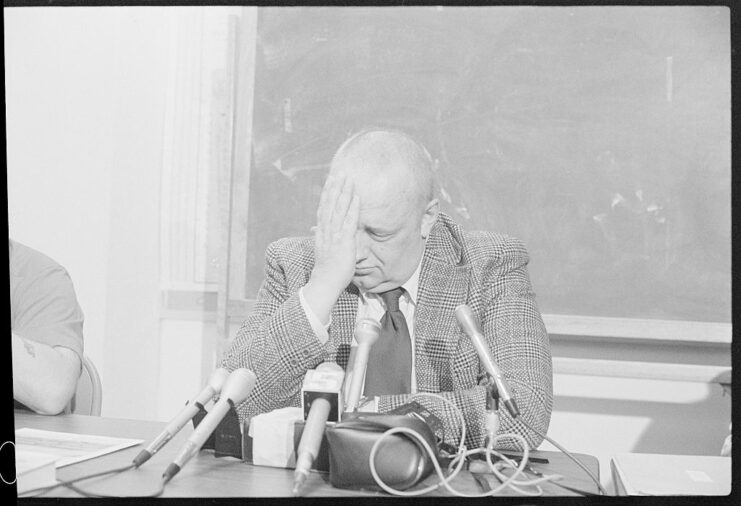 Orville Kelly holding his head in his hand while sitting before a bunch of microphones