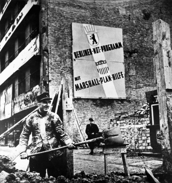 Two German construction workers standing beneath a sign promoting the Marshall Plan